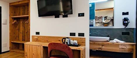 A hotel room with a wooden desk, a red chair, a bed, a TV mounted on the wall, and a bathroom with a large mirror.