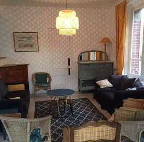 Una sala de estar acogedora con papel tapiz estampado, un aparador de madera, un sofá negro, un sillón de mimbre y una alfombra azul estampada.