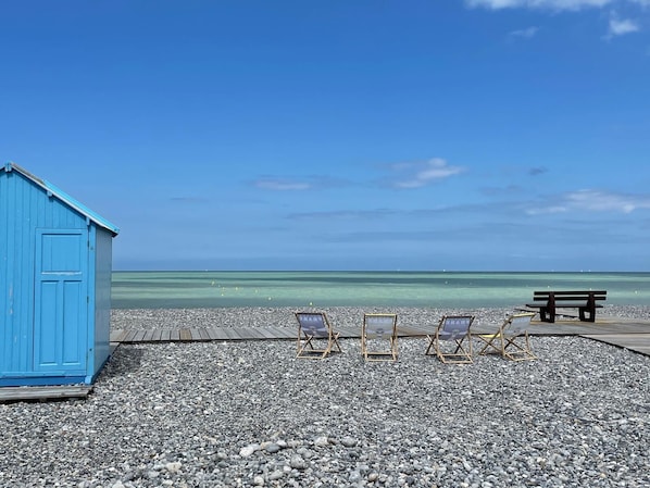 Una playa con una estructura azul, una terraza de madera y tres sillas de madera mirando al mar.