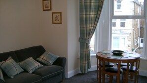 A living room with a sofa, a dining table with chairs, a window with curtains, and two framed pictures on the wall.