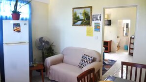 A living room with a beige sofa, a small table, a fan, and a refrigerator. There is a window with blue curtains and a potted plant on top of the refrigerator. - Louislyn Apartments #8 Quiet and Scenic (Picard)