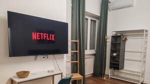A modern living room with a flat-screen TV displaying 'NETFLIX', a wooden chair, a small table with a basket, a radiator, and a white shelving unit. - Family apartment near the Vatican. Well connected. 2 terraces. WIFI. (Roma)