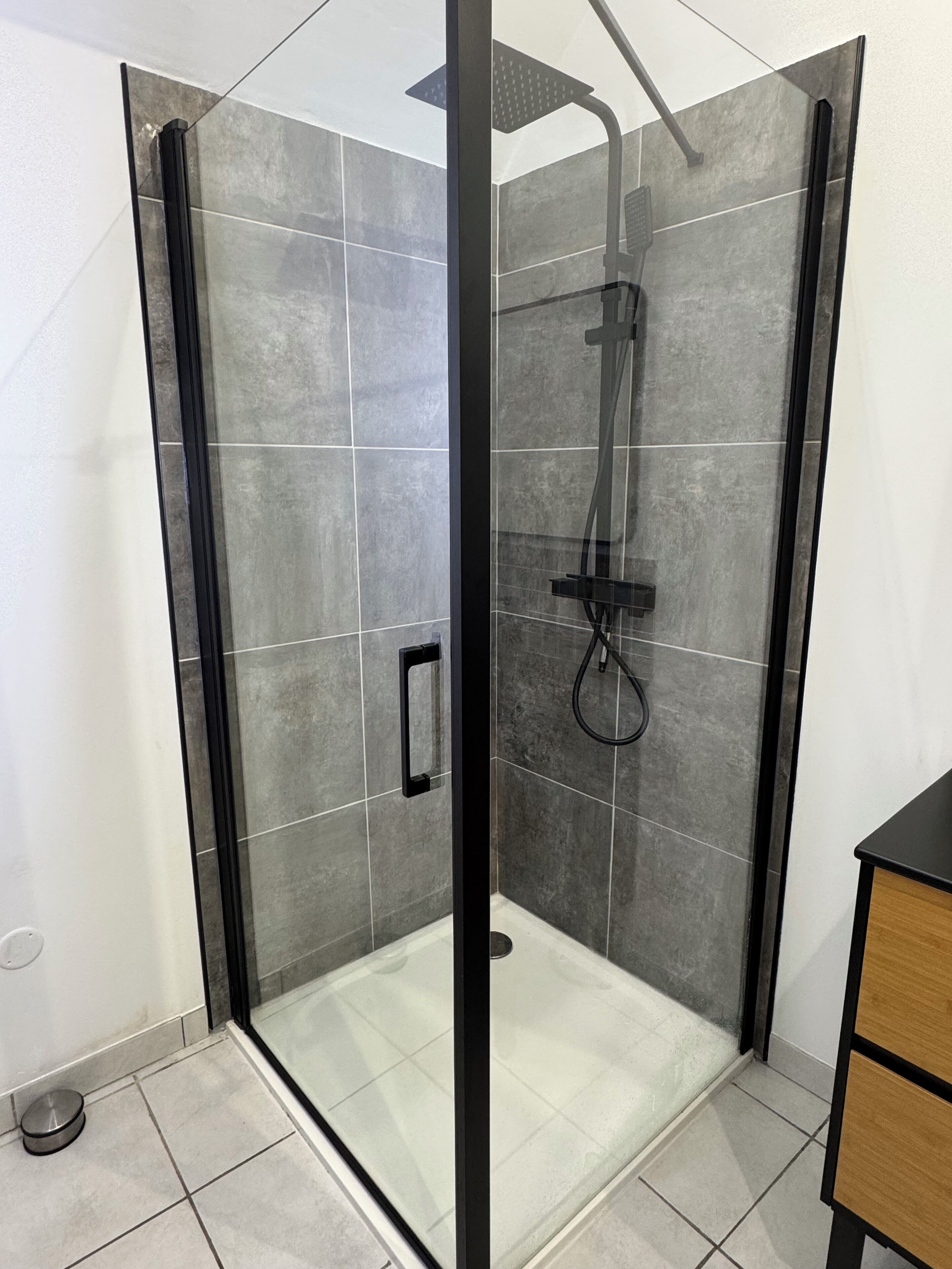 A modern shower enclosure with a glass door, grey tiles, and a black frame.