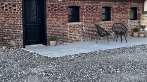 Un bâtiment en briques surmonté d’un toit en tuiles, avec une porte noire et un petit patio comprenant deux chaises et des plantes en pot.