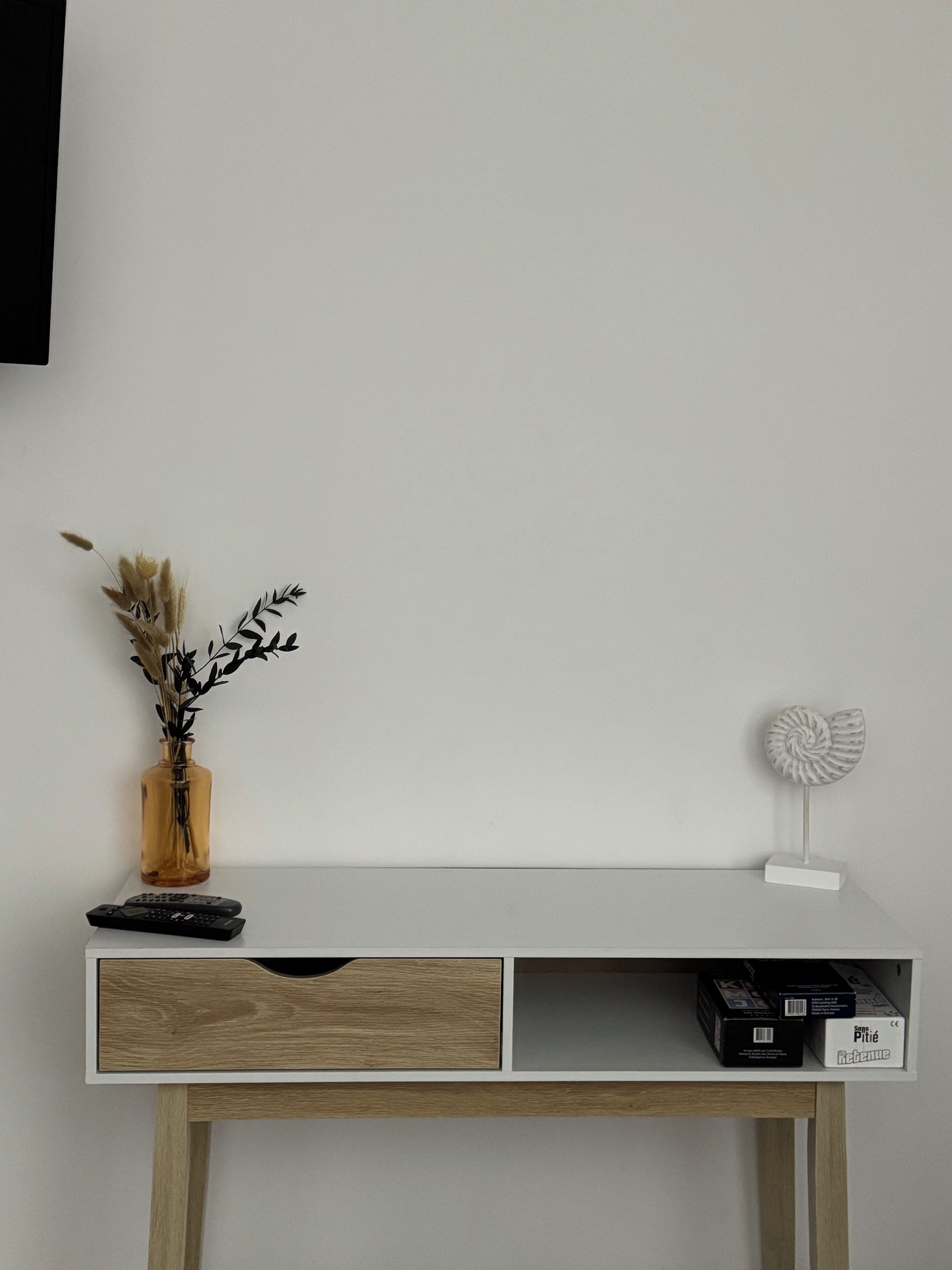 Una scrivania bianca con un cassetto, un vaso con piante essiccate, un telecomando e una conchiglia decorativa.