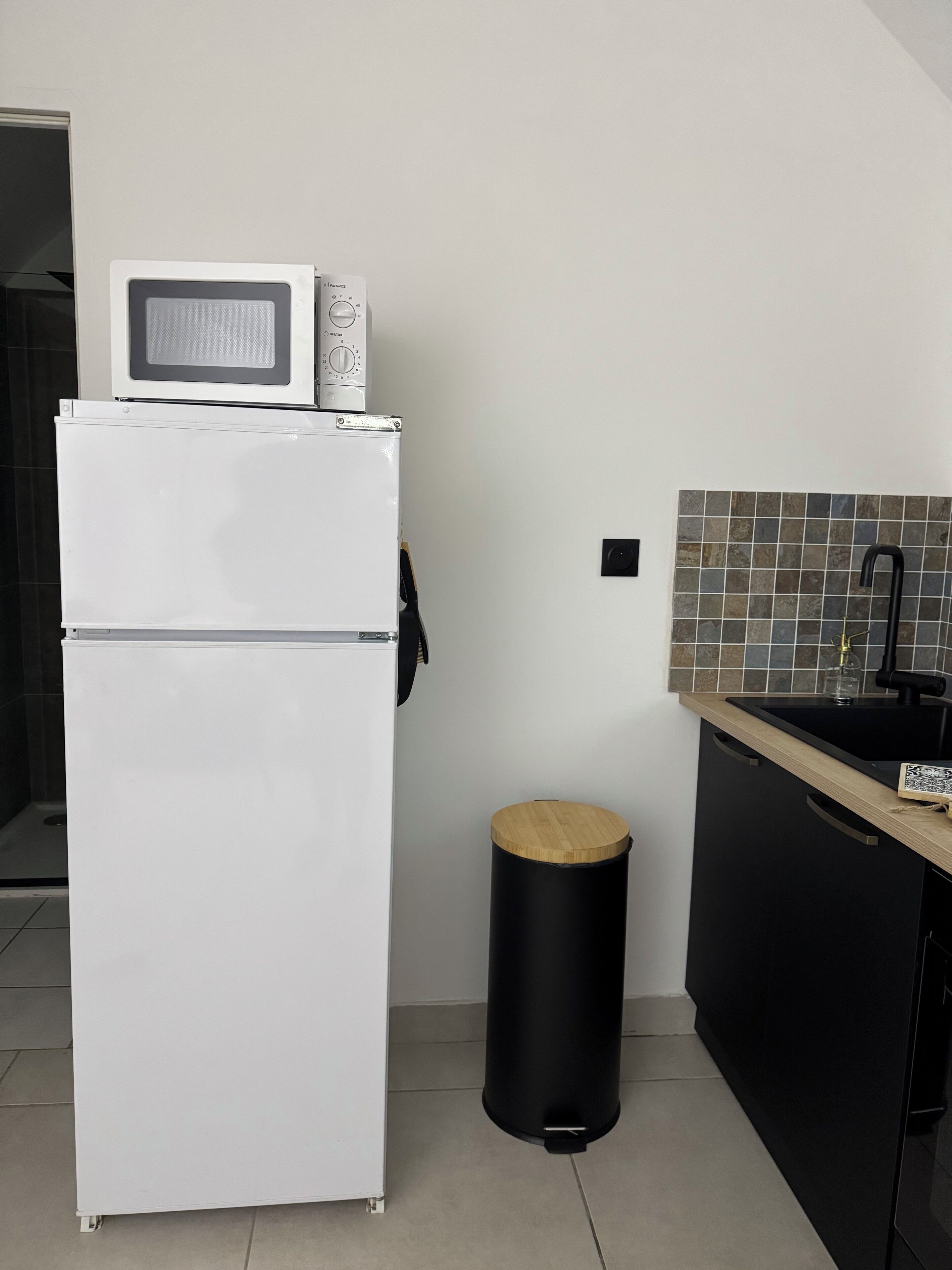 A compact kitchen with a white refrigerator, a microwave, a black trash bin, and a black sink with a tiled backsplash.