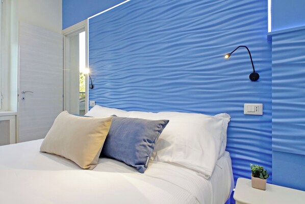 A neatly made bed with two pillows, a bedside table with a plant, and a wall-mounted light fixture. - Deluxe Cobalto Apartment - Domus Augustea San Giovanni Sunset Suites (Roma)