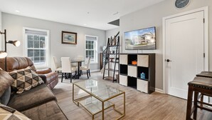 Ein modernes Wohnzimmer mit einer Ledersofa, einem Glastisch, einer Essecke mit weißem Tisch und Stühlen, einer Treppe, einem an der Wand montierten Fernseher und einer Tür mit einer Uhr.
