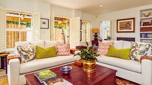 A living room with a white sofa, green and orange patterned pillows, a wooden coffee table with a plant, and a bookshelf with books and decorative items.