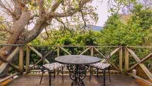 A wooden deck with a round table and four chairs, surrounded by a wooden railing and lush greenery.