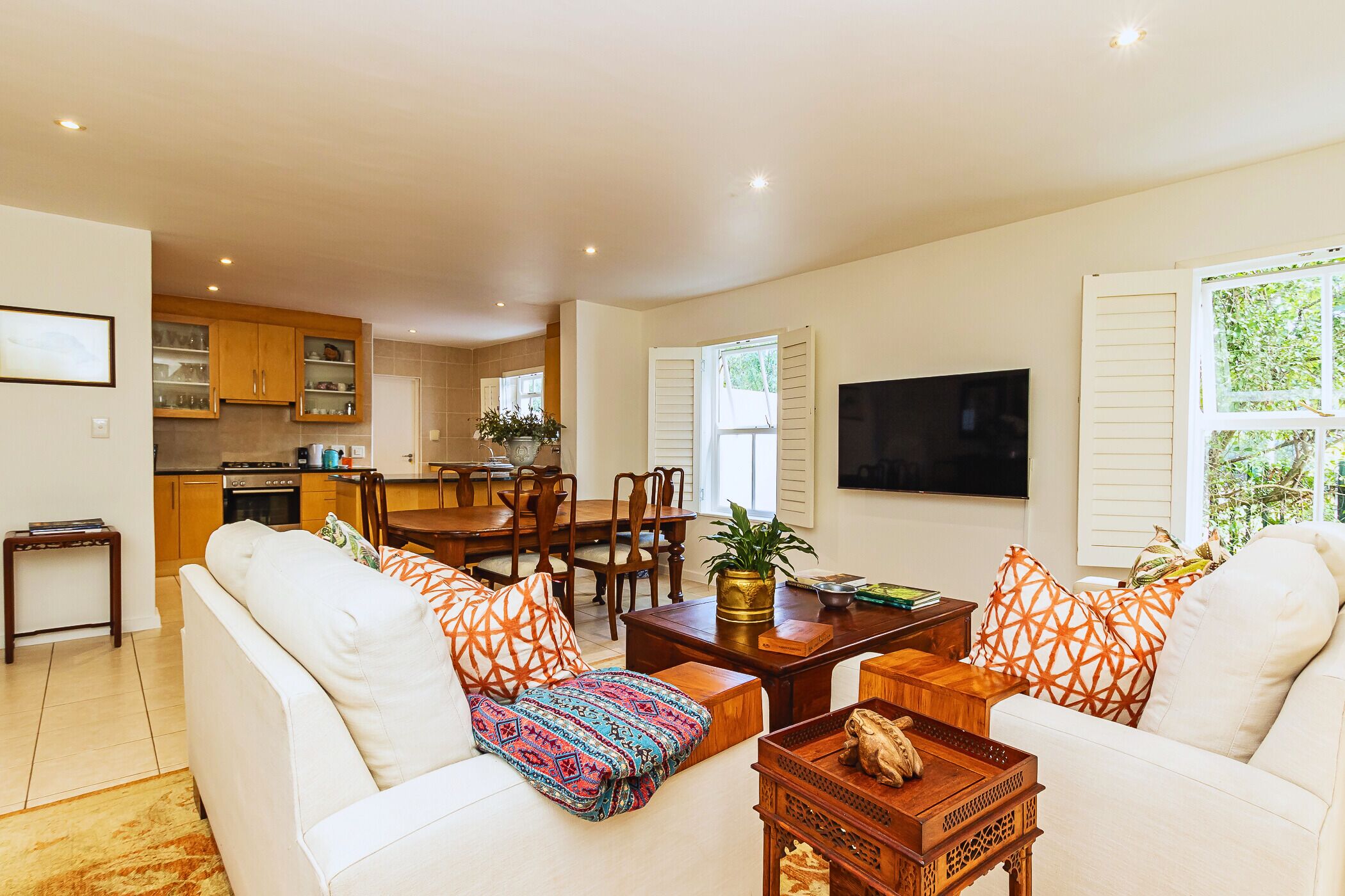 A modern living room with a sofa, coffee table, and a TV. There is a kitchen in the background with wooden cabinets and a dining area.