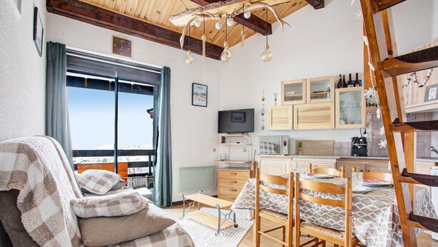 Ein gemütliches Wohnzimmer mit Sofa, Esstisch und Stühlen, einer Küchenzeile mit Schränken und einem Fernseher, sowie einem großen Fenster mit Vorhängen.