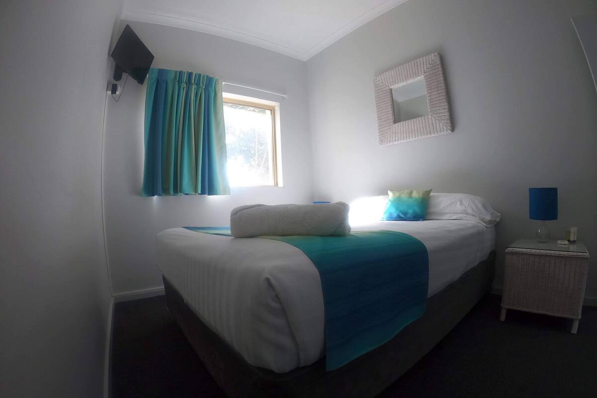 A bedroom with a bed, a nightstand, a lamp, a window with curtains, and a wall-mounted mirror.