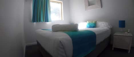 A bedroom with a bed, a nightstand, a lamp, a window with curtains, and a wall-mounted mirror.