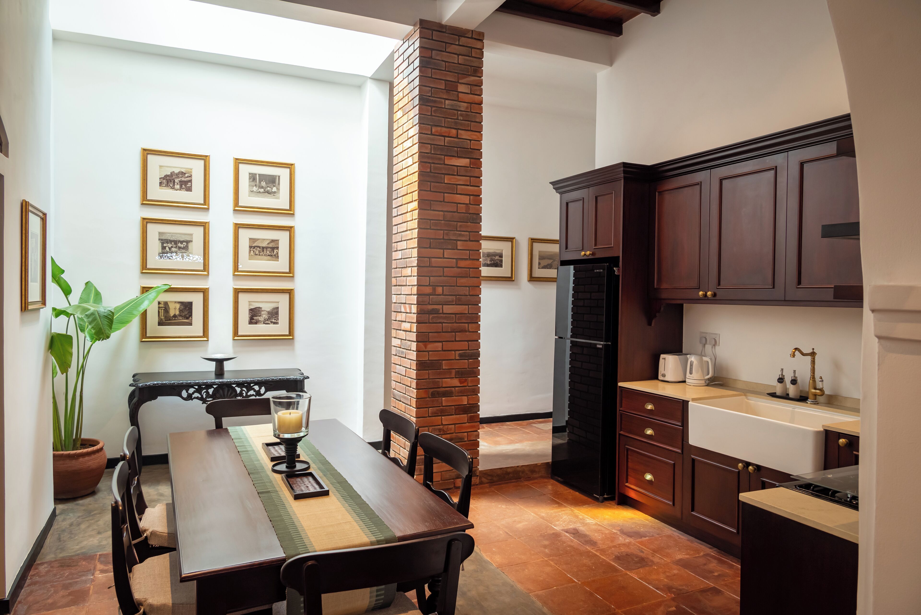 A dining area with a wooden table and chairs, a brick fireplace, framed pictures on the wall, and a kitchen with dark wooden cabinets and a sink.