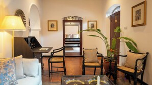Una habitación acogedora con sofá, sillas, una mesita de centro y un escritorio. Hay cuadros enmarcados en las paredes y una planta grande en la esquina.