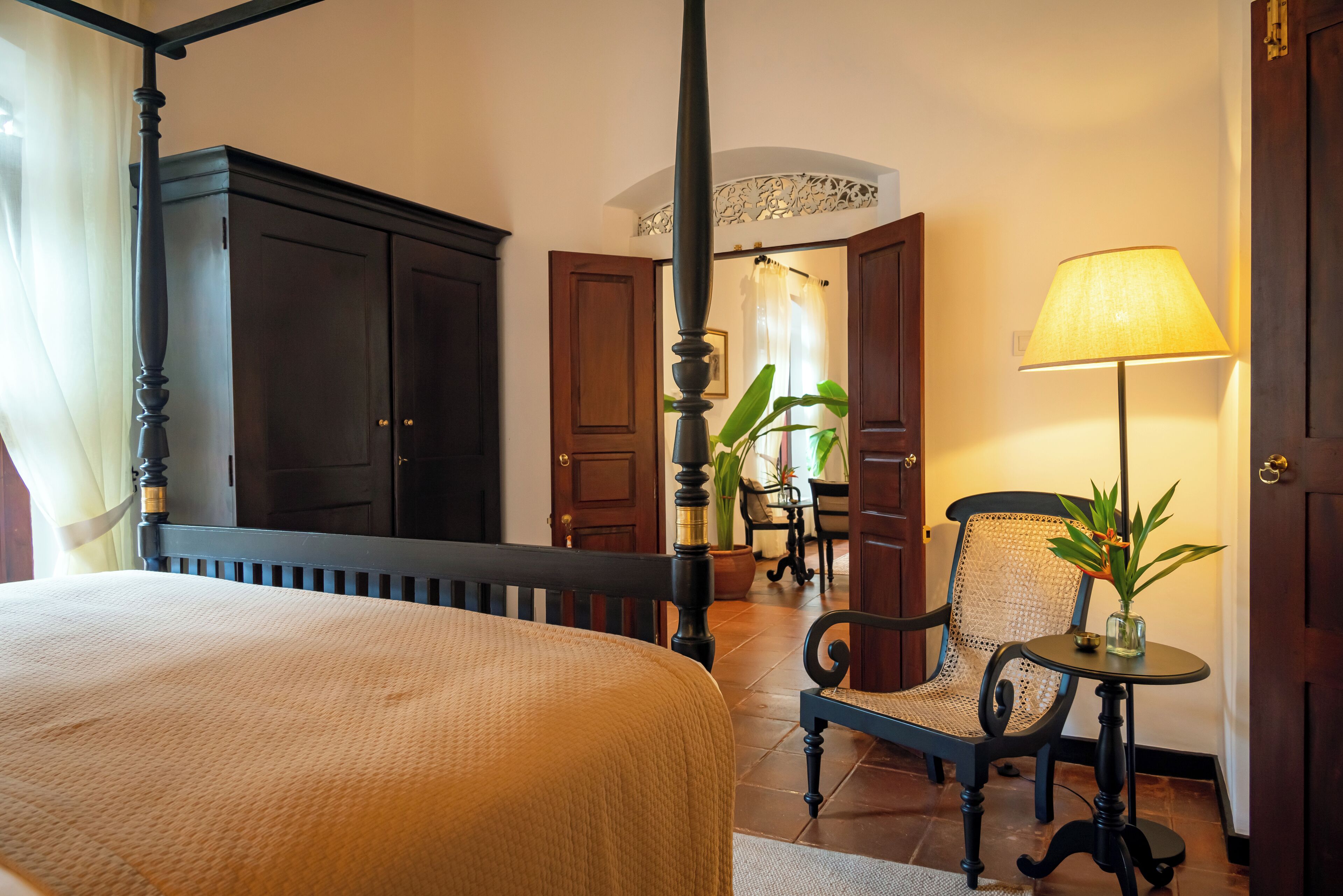 A bedroom with a four-poster bed, a wooden wardrobe, a chair, a small table with a lamp, and a potted plant.