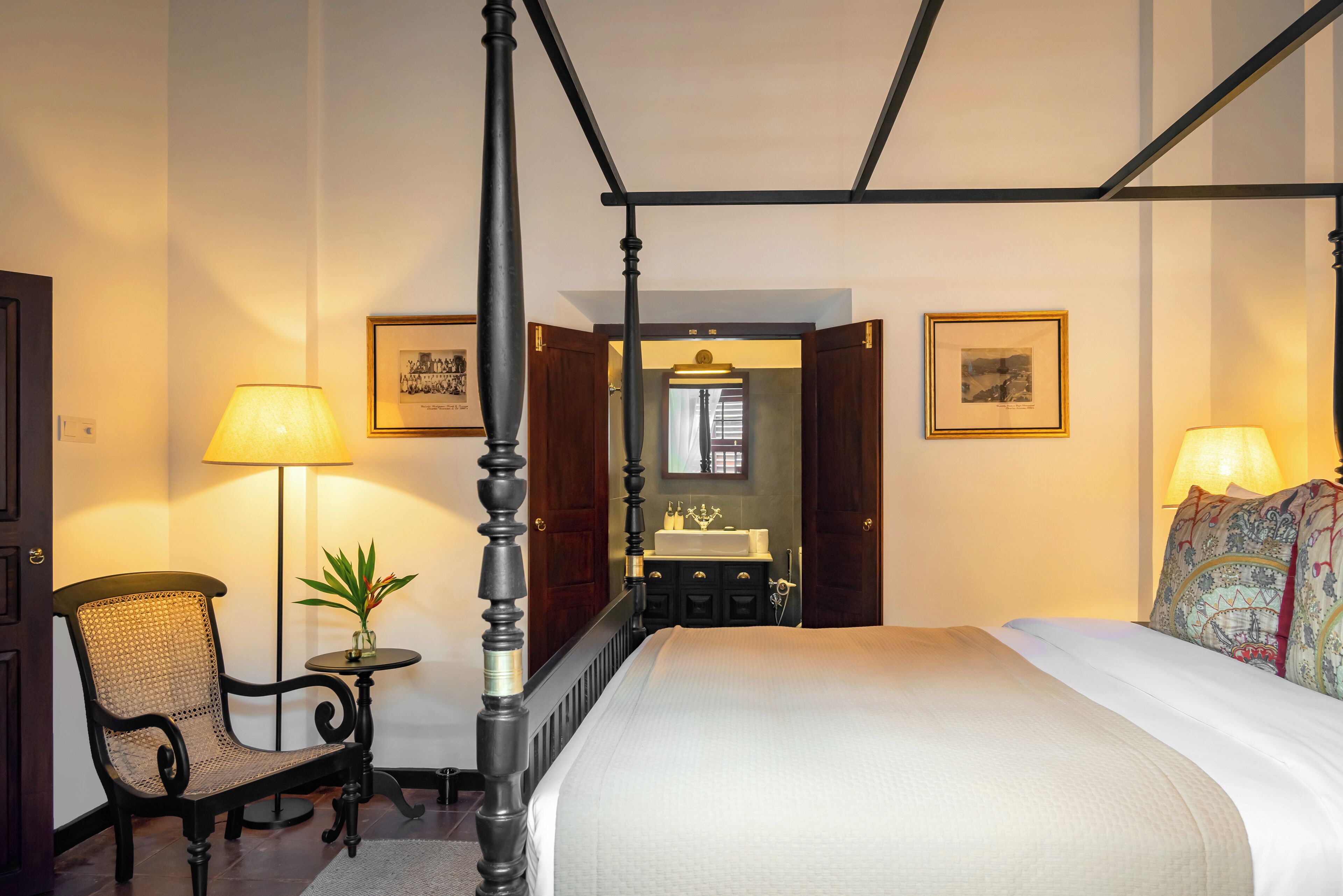Ein Bett mit Baldachin, mit weißer Bettdecke, ein Holzstuhl mit Webstuhl-Rückenlehne, ein kleiner runder Tisch mit Lampe und ein durch eine offene Tür erblicktes Badezimmer.