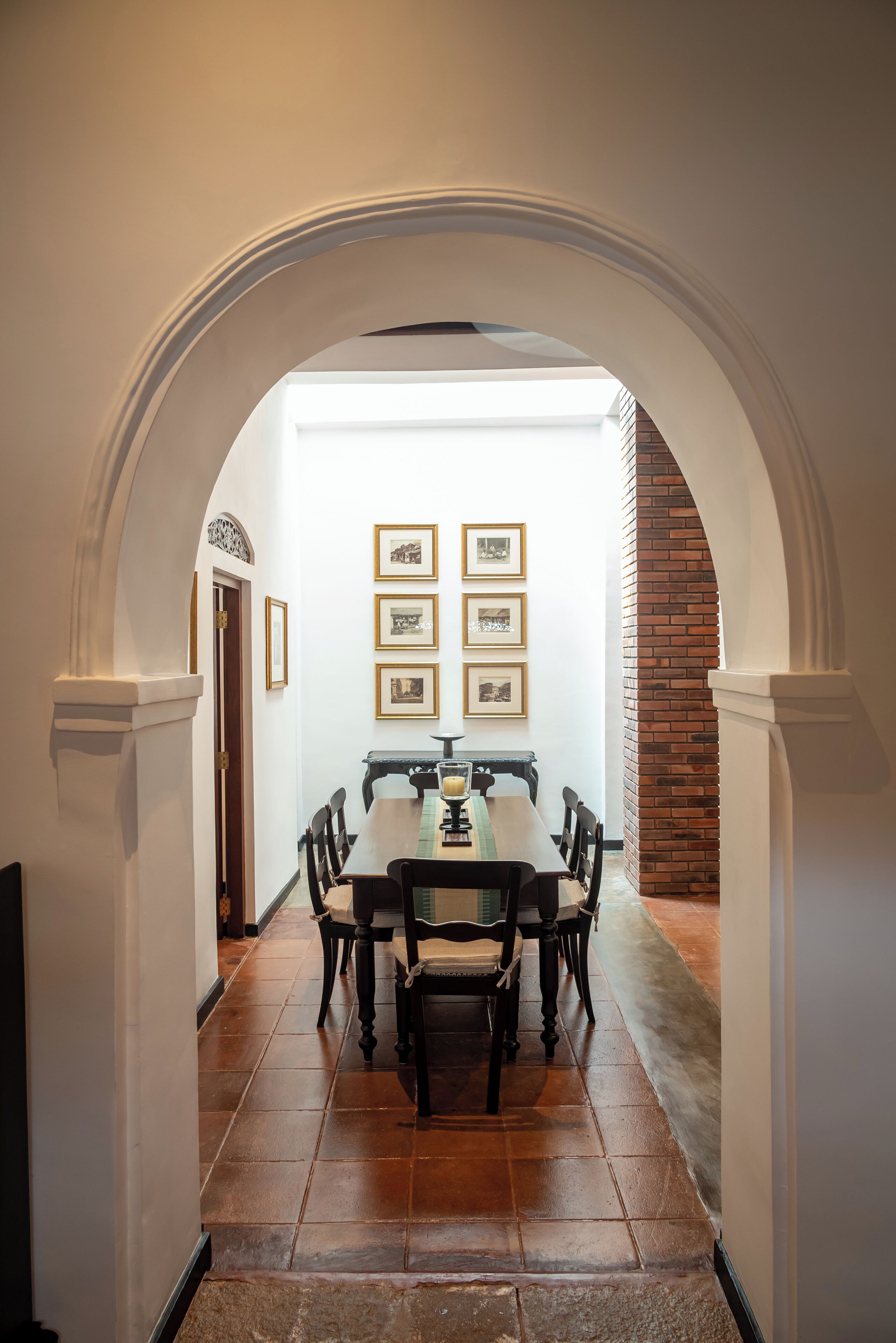 A dining area with a long table and chairs, framed pictures on the wall, and an arched doorway leading to another room.