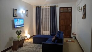 Una sala de estar con un sofá azul, un televisor montado en la pared, una puerta de madera y una ventana con cortinas.
