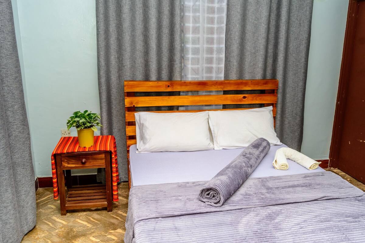 A neatly made bed with a wooden headboard, a small wooden nightstand with a potted plant, and a grey striped blanket.