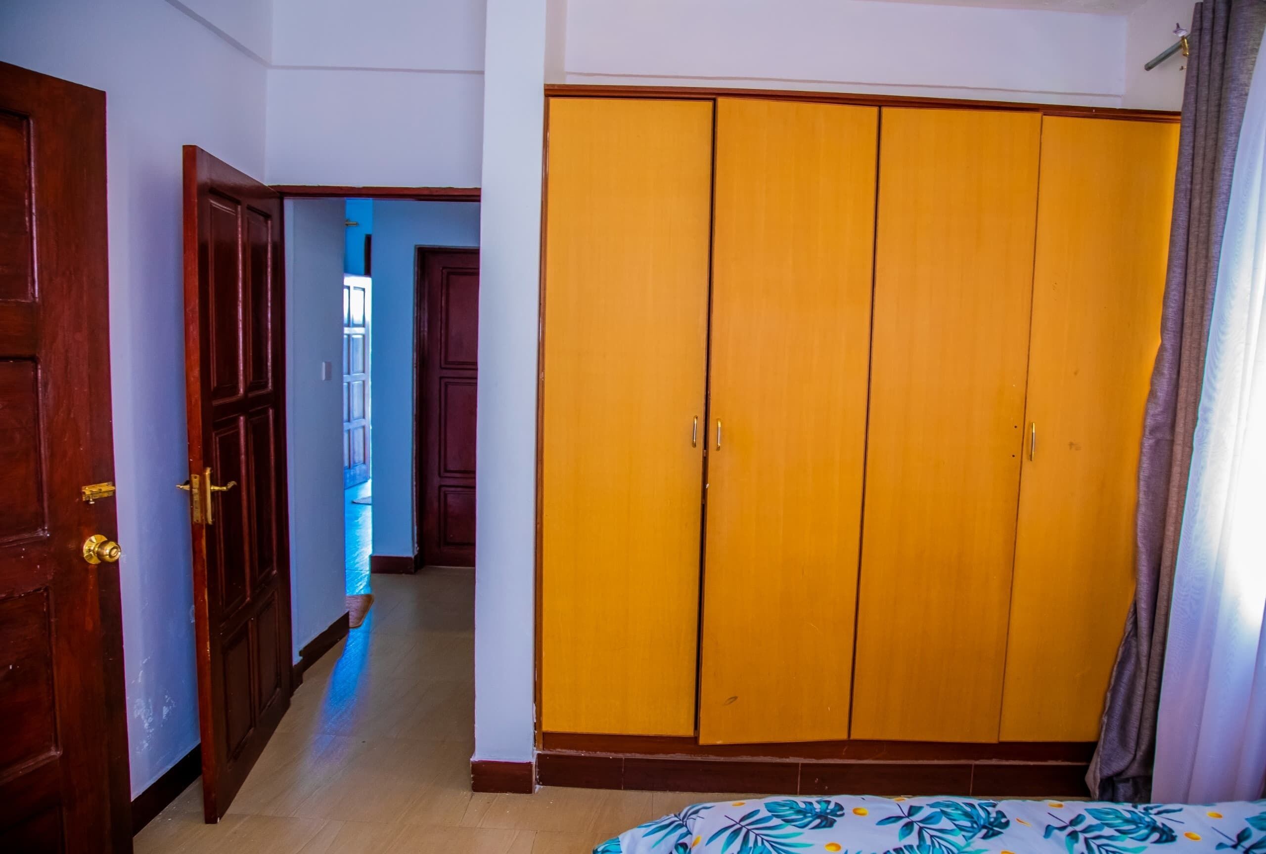 A room with a wooden wardrobe, a bed with a floral bedspread, and a view of a blue hallway.