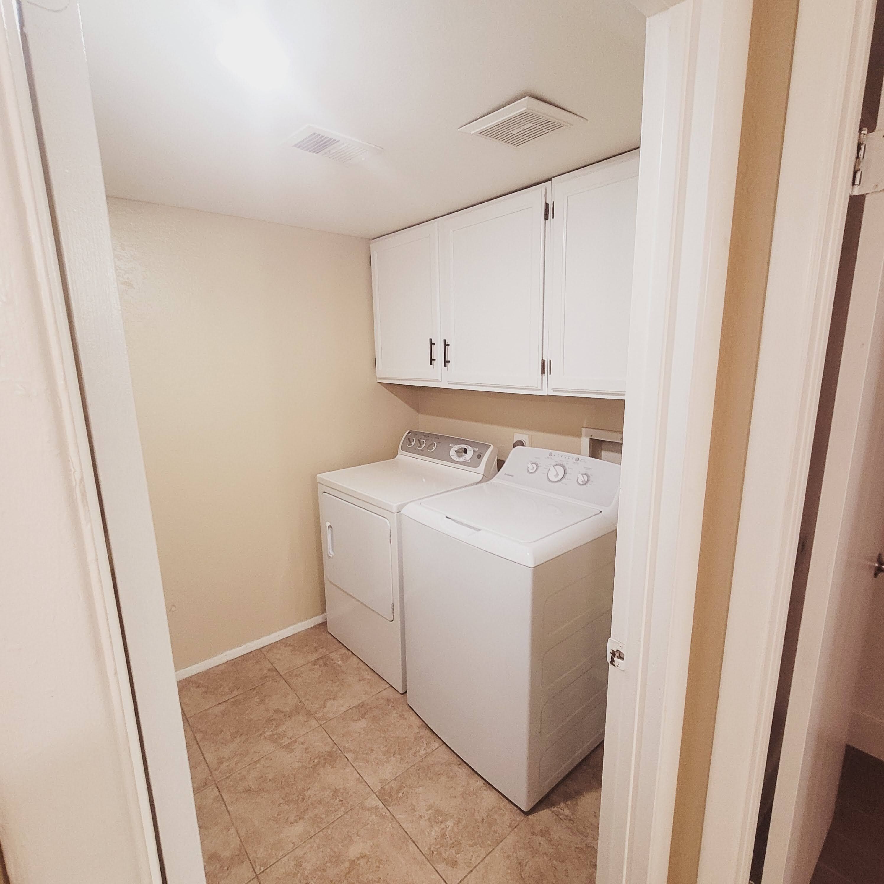 A laundry room with two white washers and dryers, beige walls, and a tiled floor.