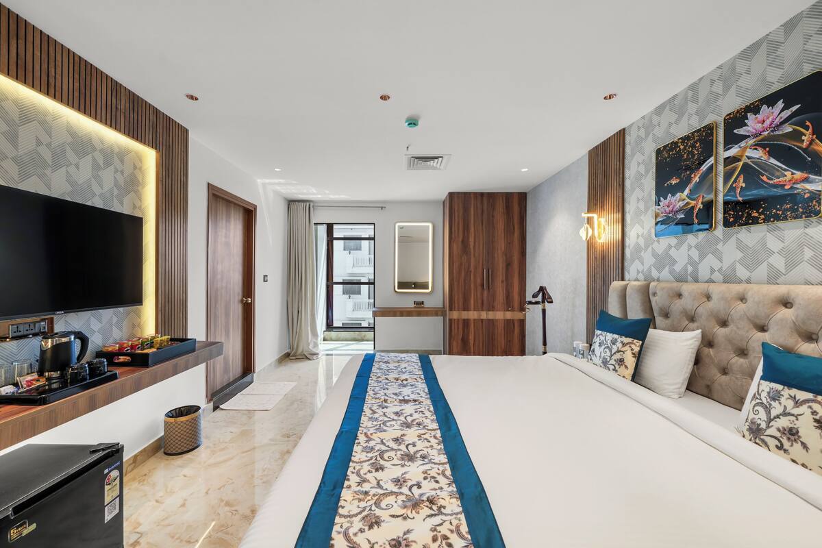A modern hotel room with a large bed, a flat-screen TV, a wooden wardrobe, and a bathroom visible through an open door.