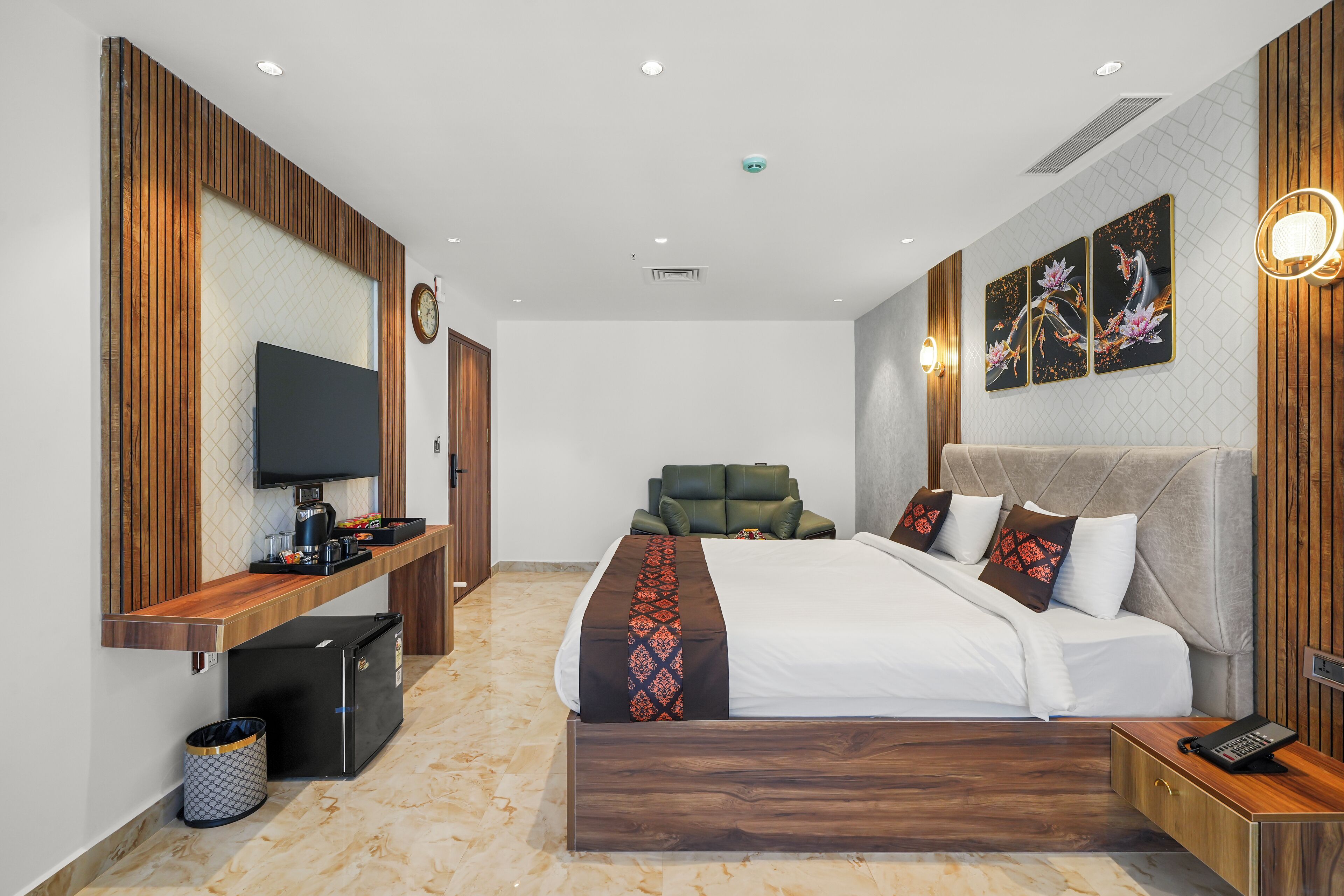 A modern hotel room with a wooden accent wall, a flat-screen TV, a sofa, a bed with patterned bedding, and artwork on the wall.