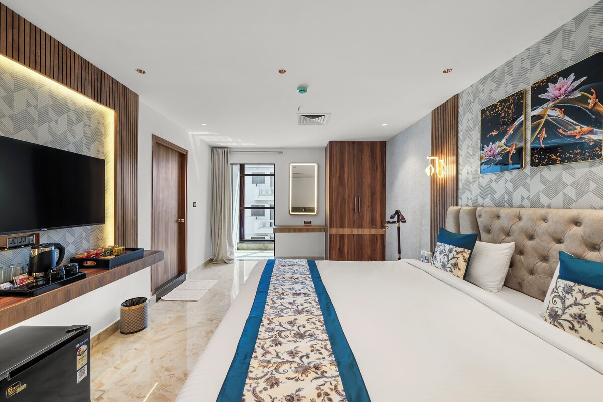 A modern hotel room with a large bed, a flat-screen TV, a wooden wardrobe, and a bathroom visible through an open door.