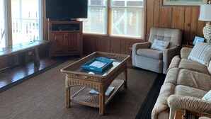 Una sala de estar con acabados de madera, un ventilador de techo, un televisor, una mesa de centro de mimbre, un sofá color beige y un cuadro enmarcado en la pared.