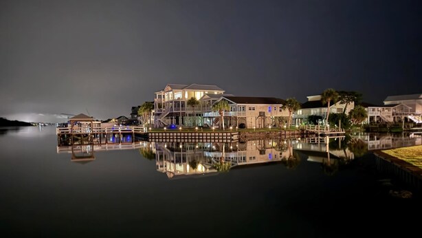 Una propiedad frente al agua con una casa grande, un muelle y un lago tranquilo que refleja los edificios.