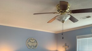 Un dormitorio con una cama, dos mesitas de noche, un ventilador de techo, un reloj de pared, un adorno en la pared y una ventana con persianas, además de una cruz en la pared.