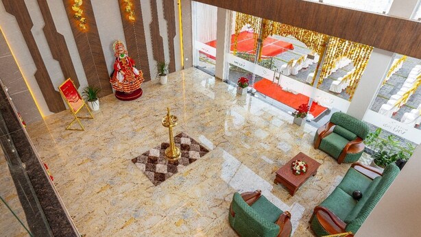A hotel lobby with a marble floor, green upholstered sofas, a central decorative pillar, and a view of a red carpeted area through large windows. - Hotel VKJ Inn (Aluva)