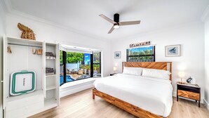 Un dormitorio con cama de madera, ventilador de techo, armario con ropa colgada y ventana con vistas a la vegetación.