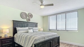 Un dormitorio con una cama de madera oscura, dos mesitas de noche con lámparas, un ventilador de techo y una ventana con persianas.