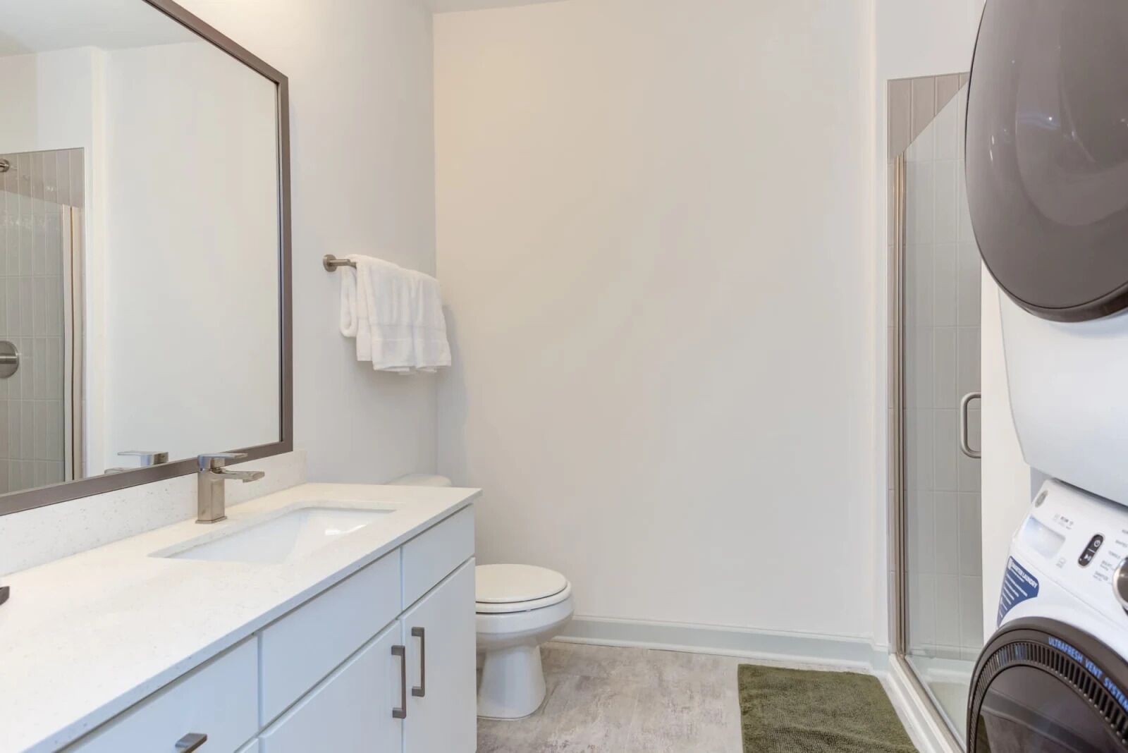 A modern bathroom with a white sink, a toilet, and a large mirror. There is a towel hanging on the wall and a hairdryer on the floor.