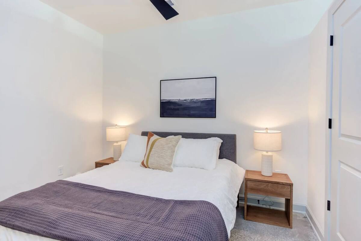 Un dormitorio ordenado con cama, dos lámparas de noche, una mesita de noche, un cuadro en la pared y una puerta.