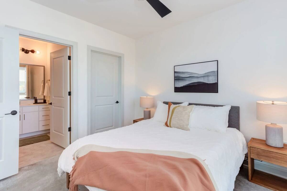 Un dormitorio con cama, lámparas de noche, una mesita de noche, una puerta que da a un baño y un cuadro en la pared.