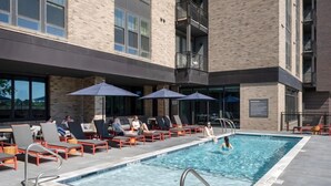 Zona de piscina con sillones de descanso y sombrillas, una piscina y fachada de edificio de ladrillo.