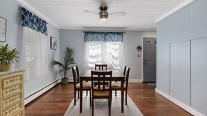 A dining room with a wooden table and chairs, a ceiling fan, and a window with patterned curtains.