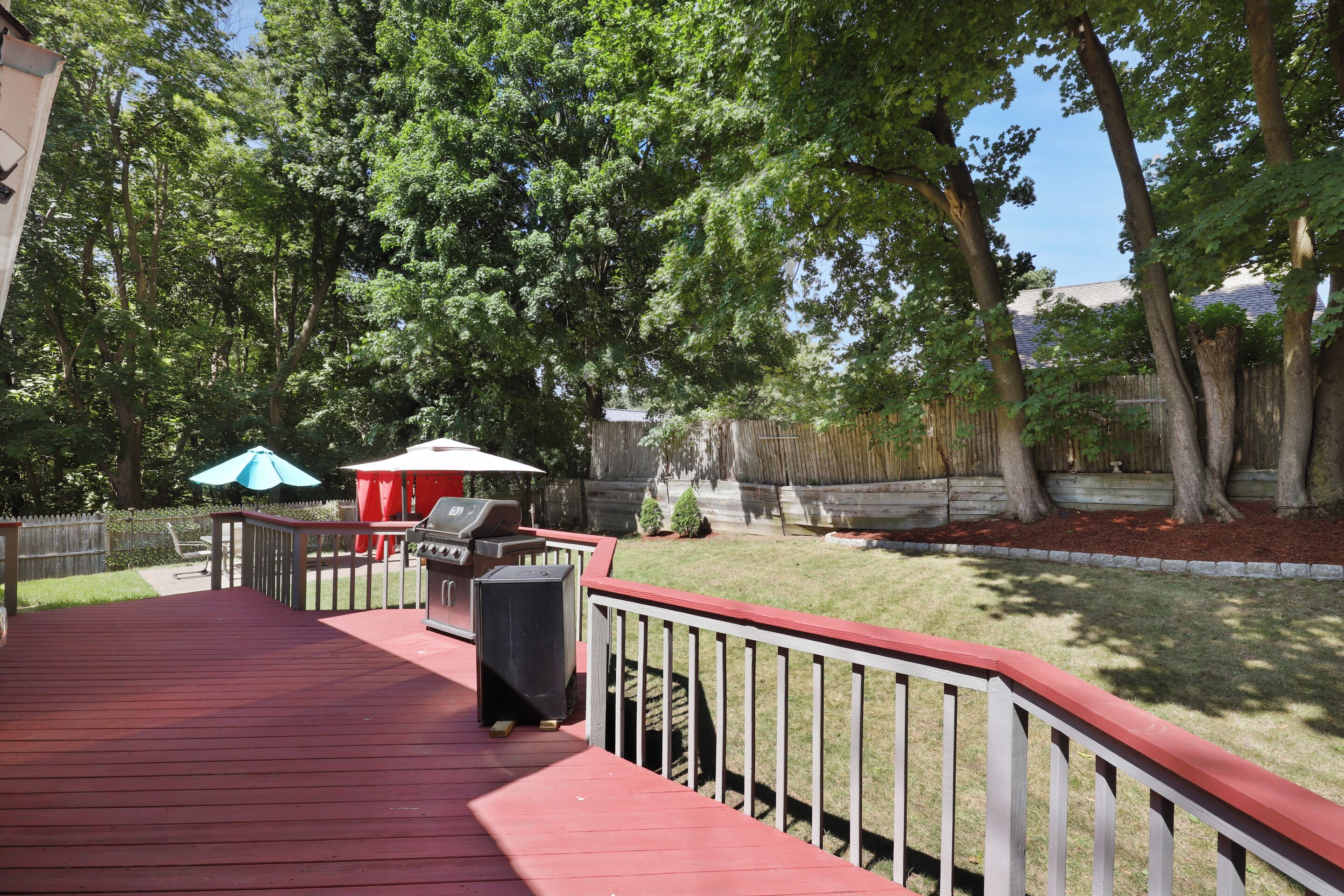 Une terrasse en bois avec une rambarde rouge, un barbecue et un parasol bleu.