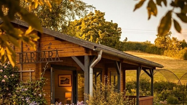 A wooden building with a covered porch, surrounded by landscaped gardens and a field.