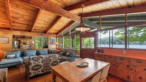 A cozy cabin interior with a wooden dining table, a sofa set, and large windows offering a view of the outdoors.
