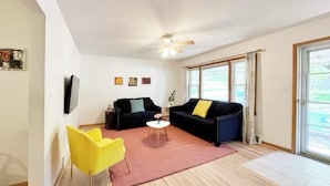 Ein Wohnzimmer mit zwei schwarzen Sofas, einem gelben Sessel, einem kleinen weißen Tisch und einem Deckenventilator.