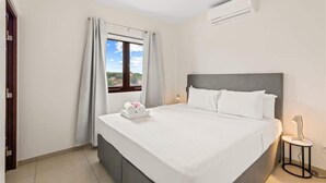 A hotel room with a bed, a window with curtains, a bedside table with a lamp, and an air conditioning unit on the wall. - Searenity View | Blue Bay Caribbean Escape (Blue Bay Golf &amp; Beach Resort)