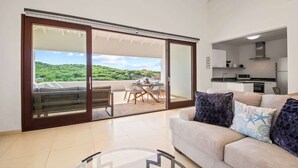 A modern living room with a sliding glass door leading to a balcony, a sofa with decorative pillows, and a kitchen area visible through an open door. - Searenity View | Blue Bay Caribbean Escape (Blue Bay Golf &amp; Beach Resort)