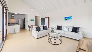 A modern living room with a white sofa, a glass coffee table, and a wooden side table. There is a kitchen visible in the background. - Searenity View | Blue Bay Caribbean Escape (Blue Bay Golf &amp; Beach Resort)