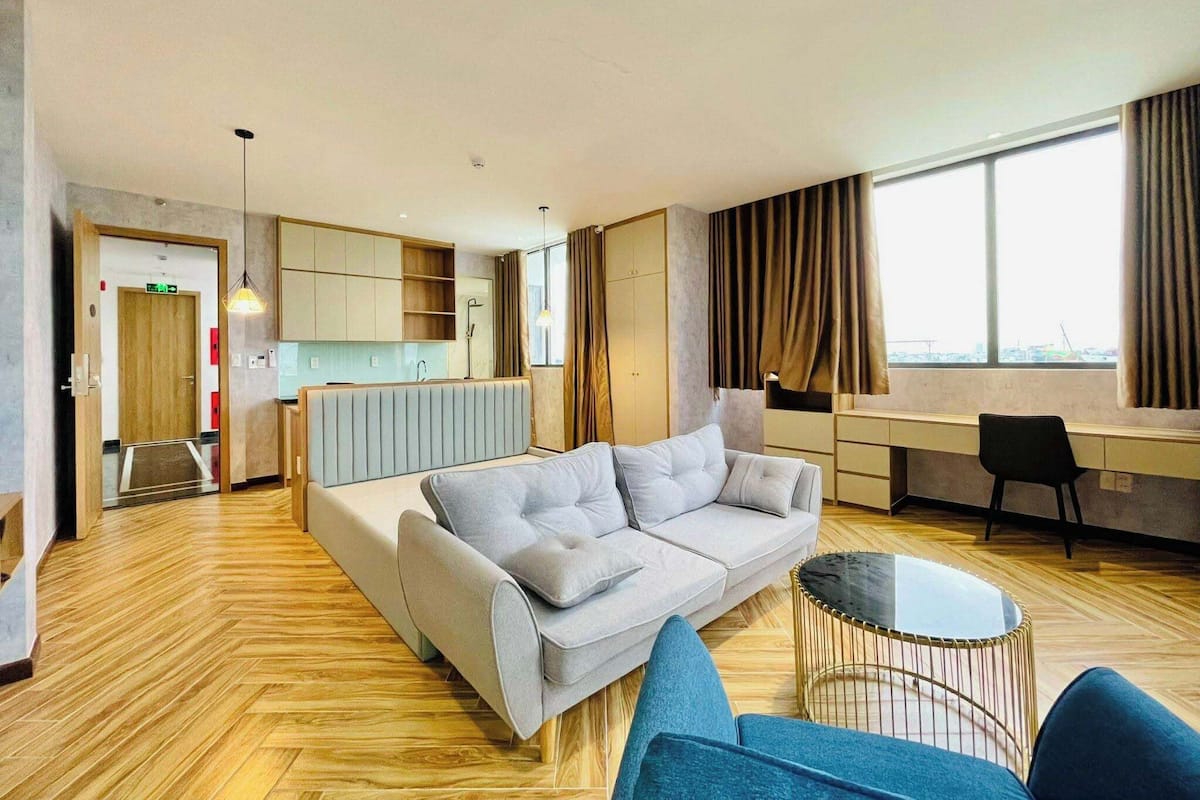 A modern hotel room with a sofa, a small round table, a desk, and a view of the city through large windows.