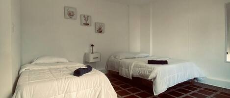 A room with two beds, a white wall, a small bedside table with a lamp, and a tiled floor.